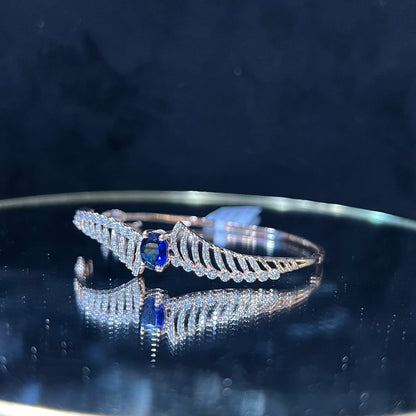 A luxurious gold diamond bracelet with a brilliant cut blue stone in an openable design, displayed on a reflective surface.