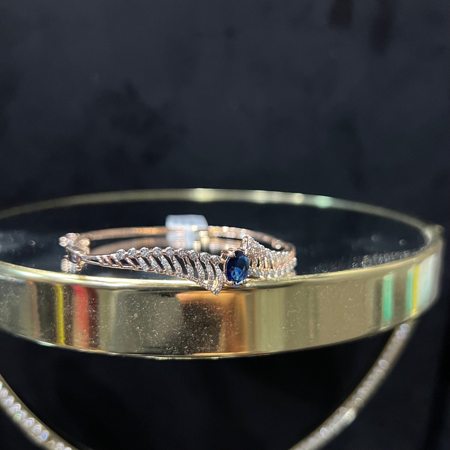 A luxurious gold diamond bracelet with a brilliant cut blue stone in an openable design, displayed on a reflective surface.