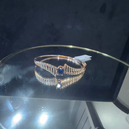 A luxurious gold diamond bracelet with a brilliant cut blue stone in an openable design, displayed on reflective surface.