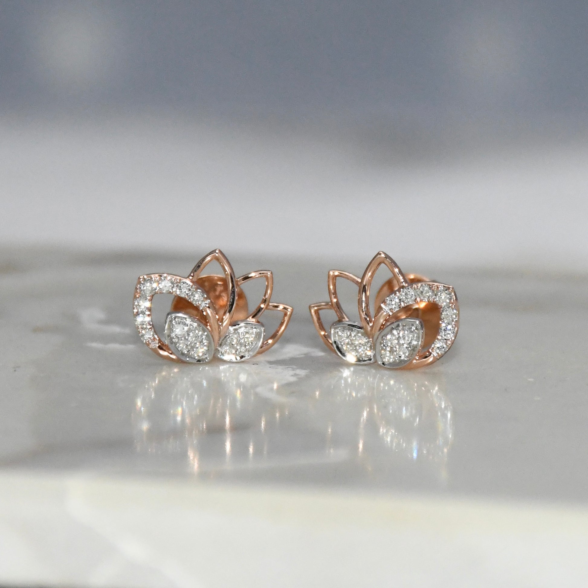 Rose gold lotus flower earrings with clear diamonds on a reflective surface.