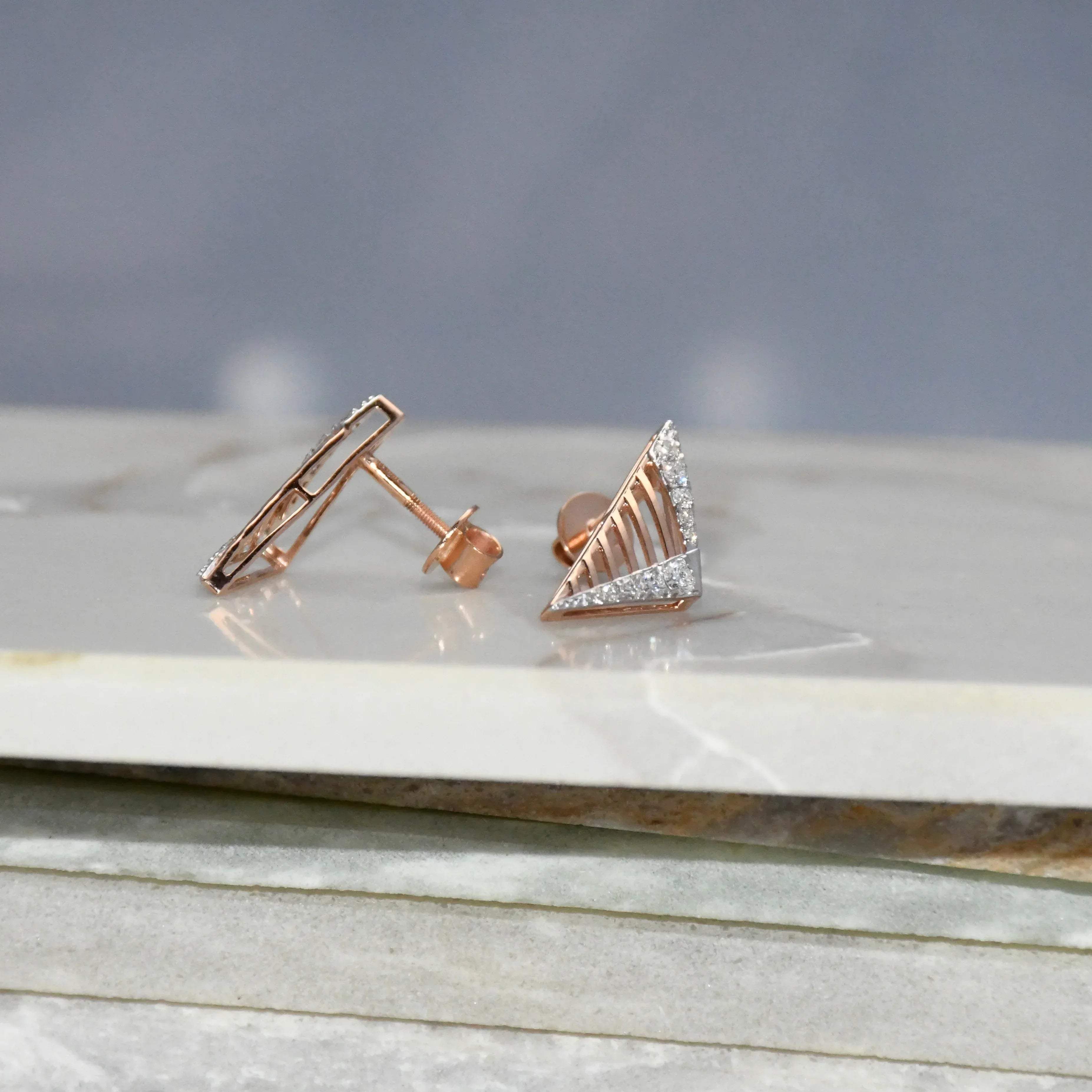 Gold triangle-shaped diamond earrings on a marble surface with a gray background.