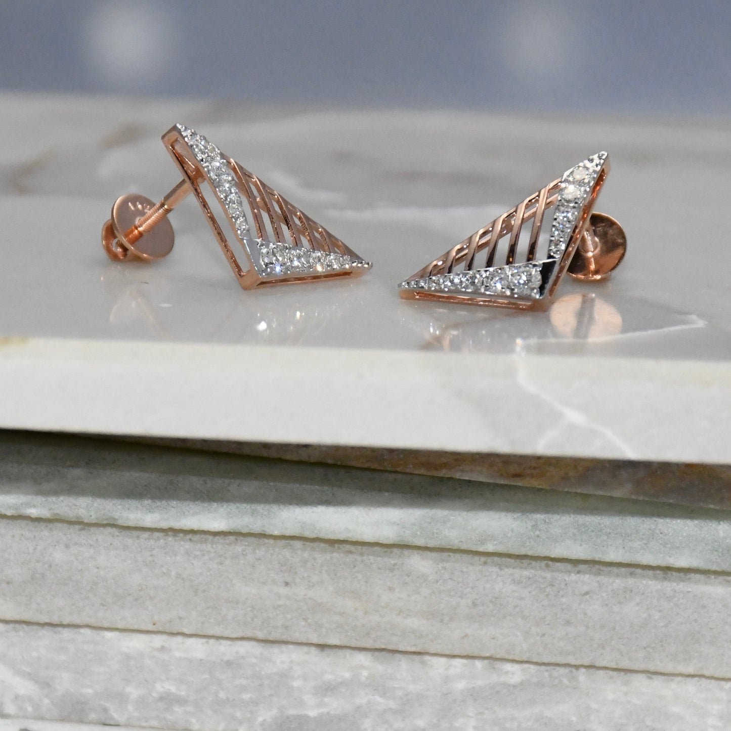 Gold triangle-shaped diamond earrings on a marble surface with a gray background.