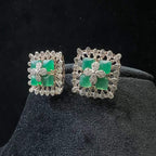 Square-shaped diamond earrings with green gemstone center and floral diamond design, placed on a black display stand.