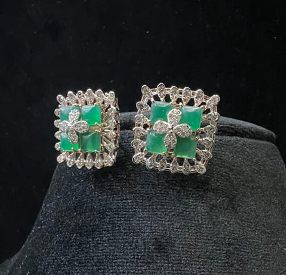 Square-shaped diamond earrings with green gemstone center and floral diamond design, placed on a black display stand.