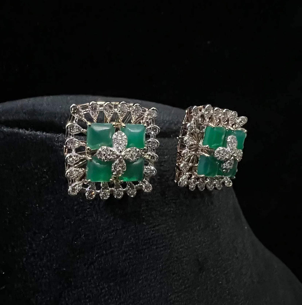 Square-shaped diamond earrings with green gemstone center and floral diamond design, placed on a black display stand.