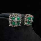 Square-shaped diamond earrings with green gemstone center and floral diamond design, placed on a black display stand.