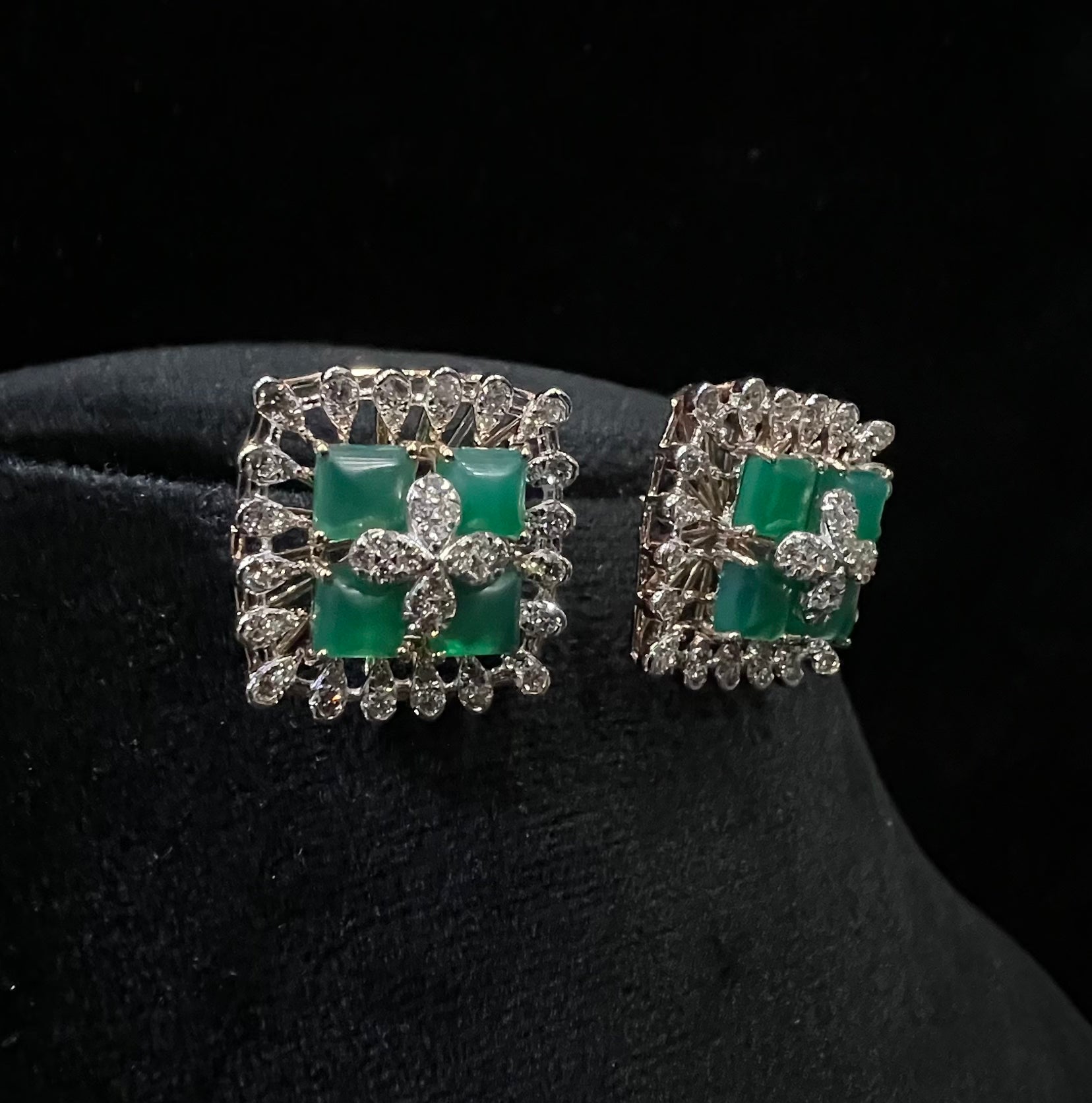 Square-shaped diamond earrings with green gemstone center and floral diamond design, placed on a black display stand.