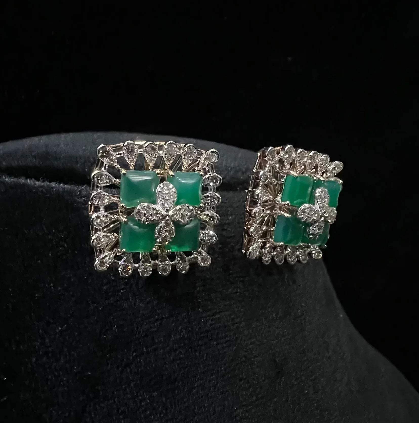 Square-shaped diamond earrings with green gemstone center and floral diamond design, placed on a black display stand.