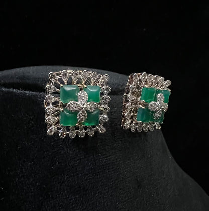 Square-shaped diamond earrings with green gemstone center and floral diamond design, placed on a black display stand.