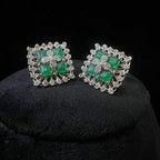 Square-shaped diamond earrings with green gemstone center and floral diamond design, placed on a black display stand.