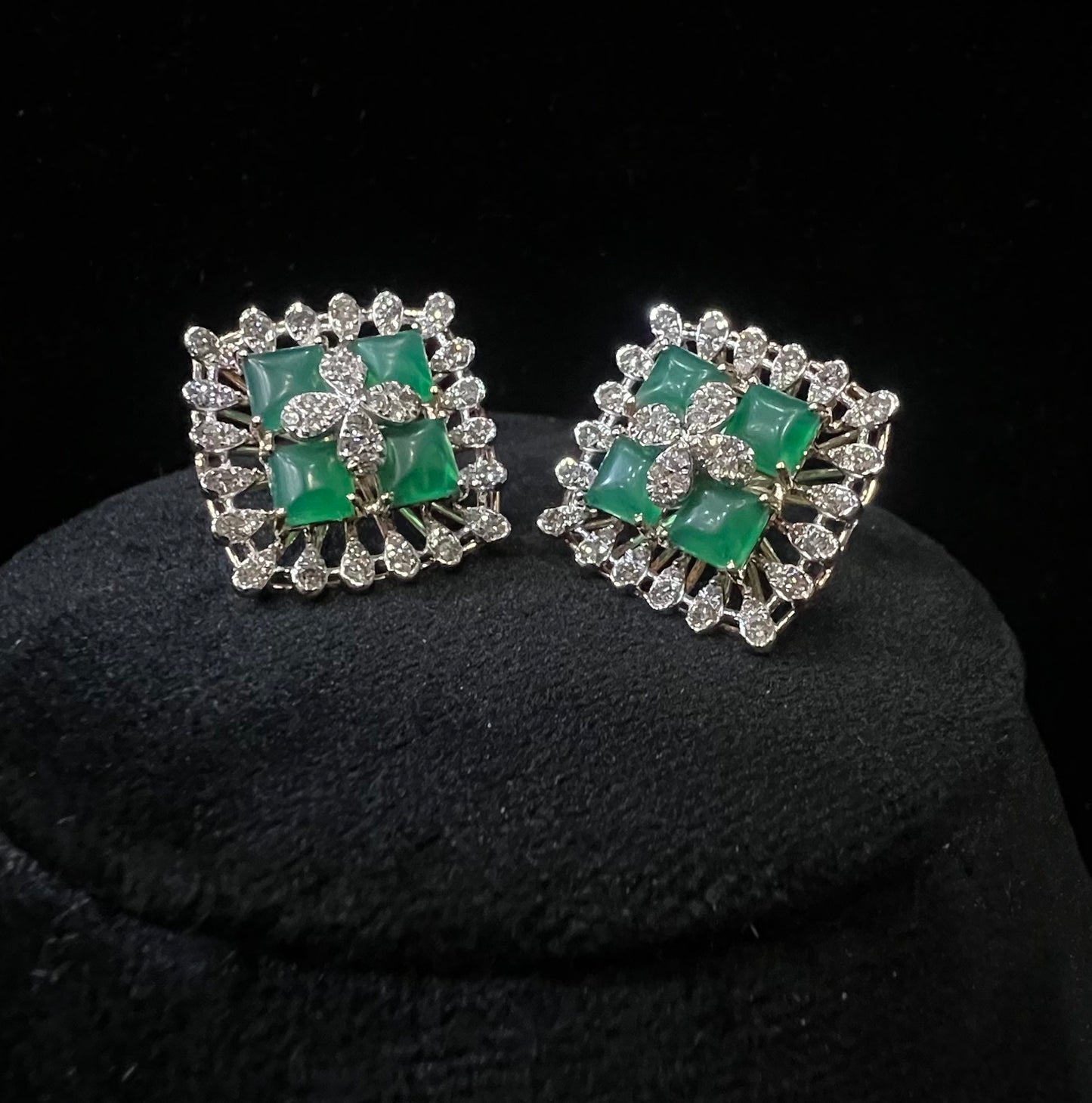 Square-shaped diamond earrings with green gemstone center and floral diamond design, placed on a black display stand.