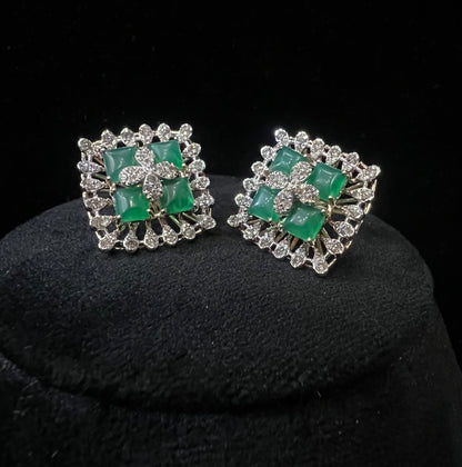 Square-shaped diamond earrings with green gemstone center and floral diamond design, placed on a black display stand.