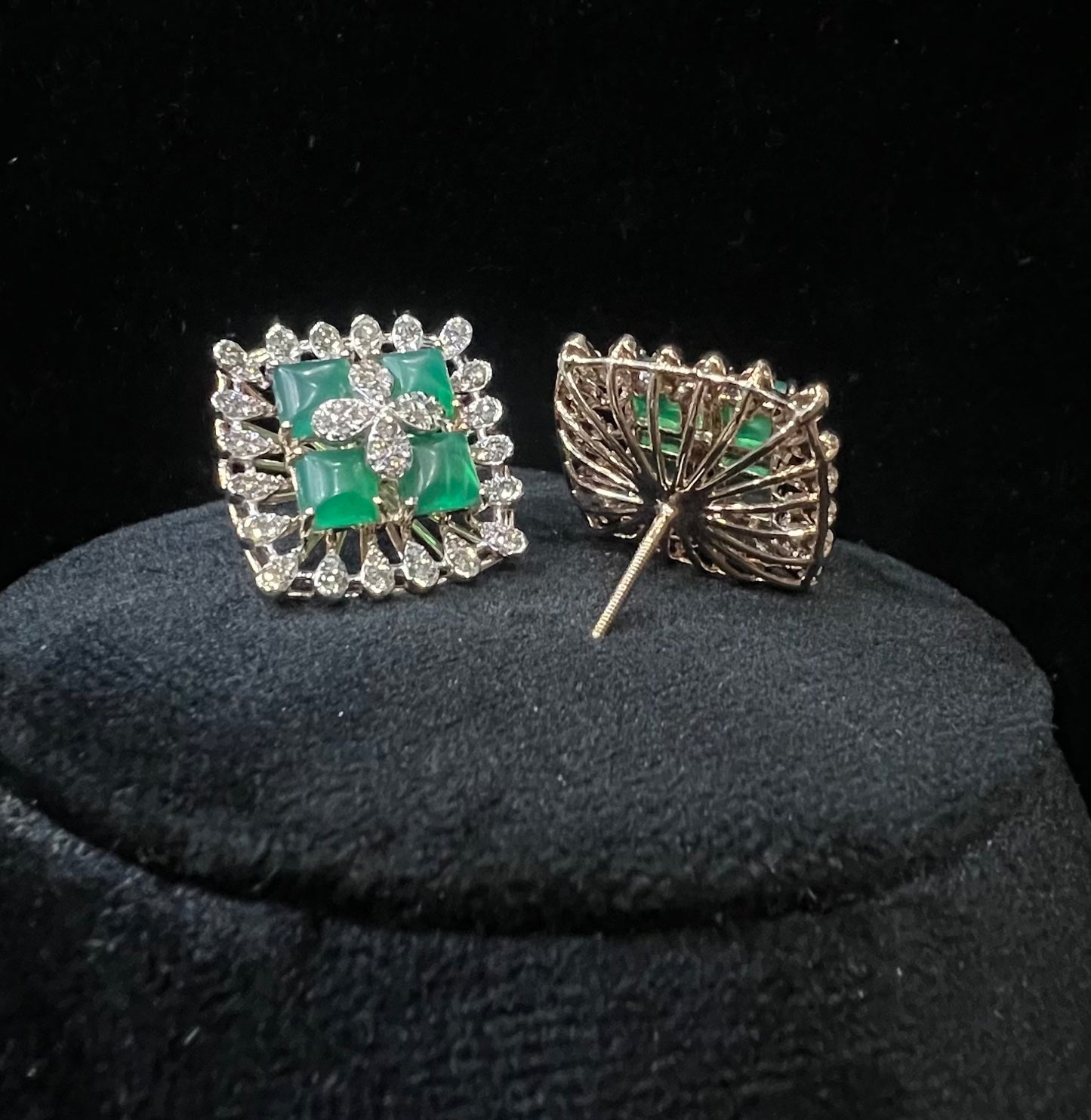 Square-shaped diamond earrings with green gemstone center and floral diamond design, placed on a black display stand.