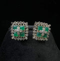 Square-shaped diamond earrings with green gemstone center and floral diamond design, placed on a black display stand.