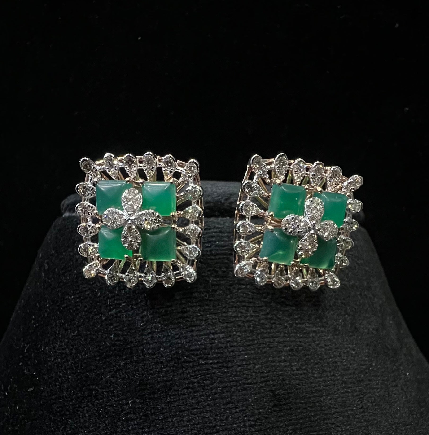 Square-shaped diamond earrings with green gemstone center and floral diamond design, placed on a black display stand.