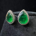 Pair of teardrop green stone and diamond halo earrings displayed on a black stand.
