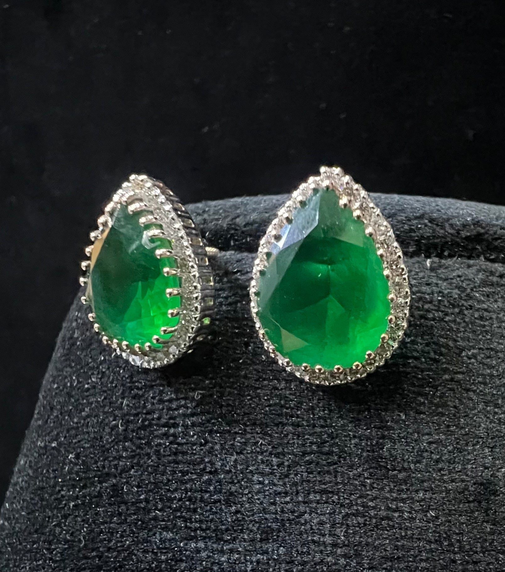 Pair of teardrop green stone and diamond halo earrings displayed on a black stand.