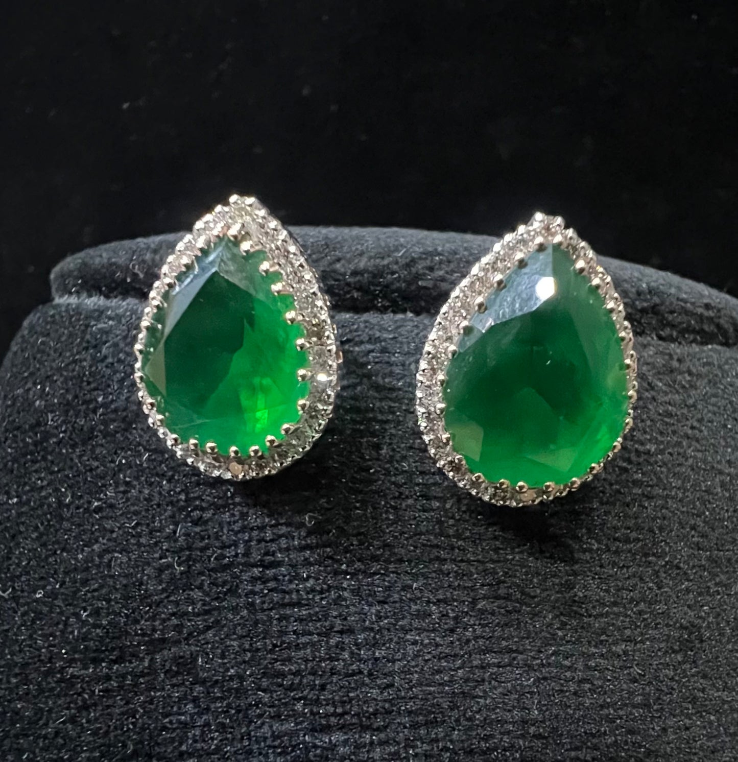 Pair of teardrop green stone and diamond halo earrings displayed on a black stand.