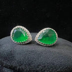 Pair of teardrop green stone and diamond halo earrings displayed on a black stand.