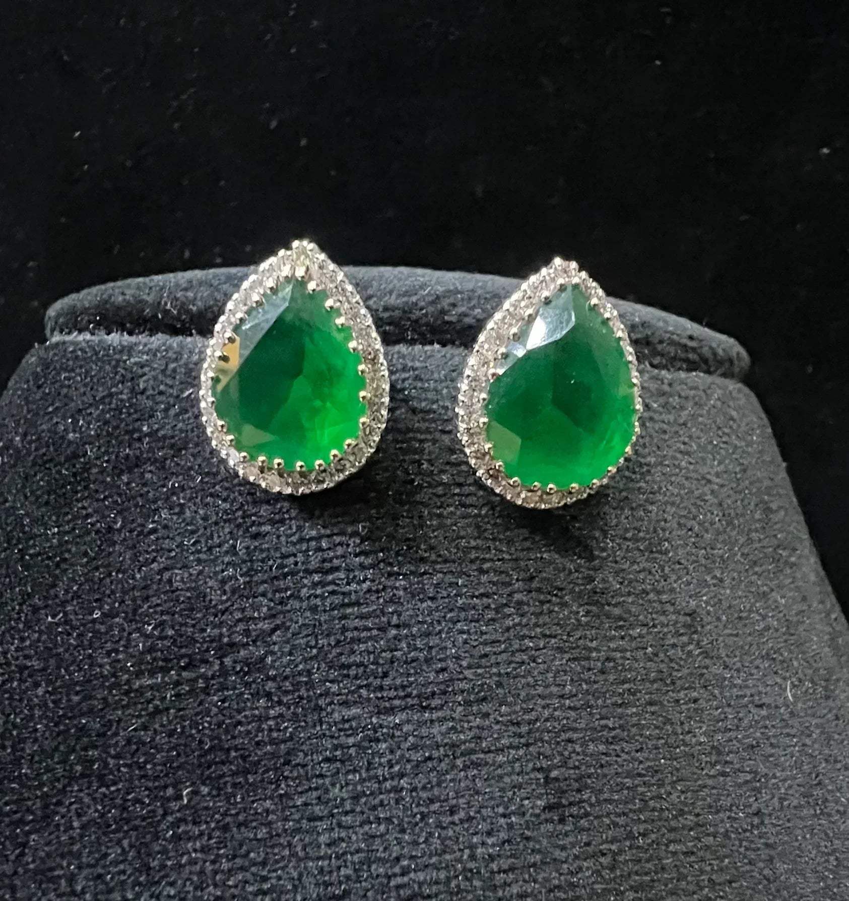 Pair of teardrop green stone and diamond halo earrings displayed on a black stand.