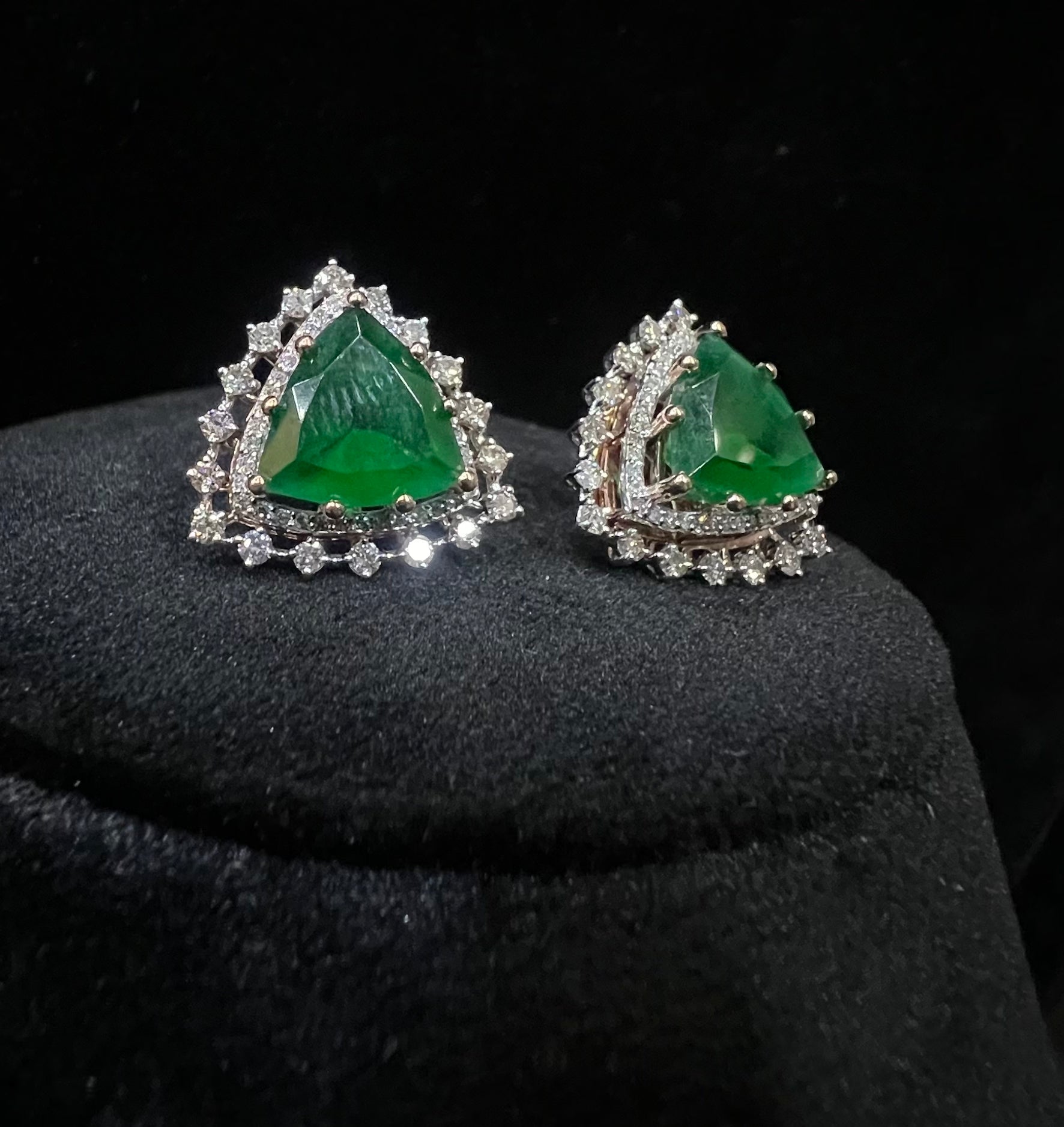 A pair of gold diamond earrings with a green color stone set in a minimalist design on a black background.