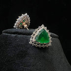 A pair of gold diamond earrings with a green color stone set in a minimalist design on a black background.