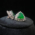 A pair of gold diamond earrings with a green color stone set in a minimalist design on a black background.