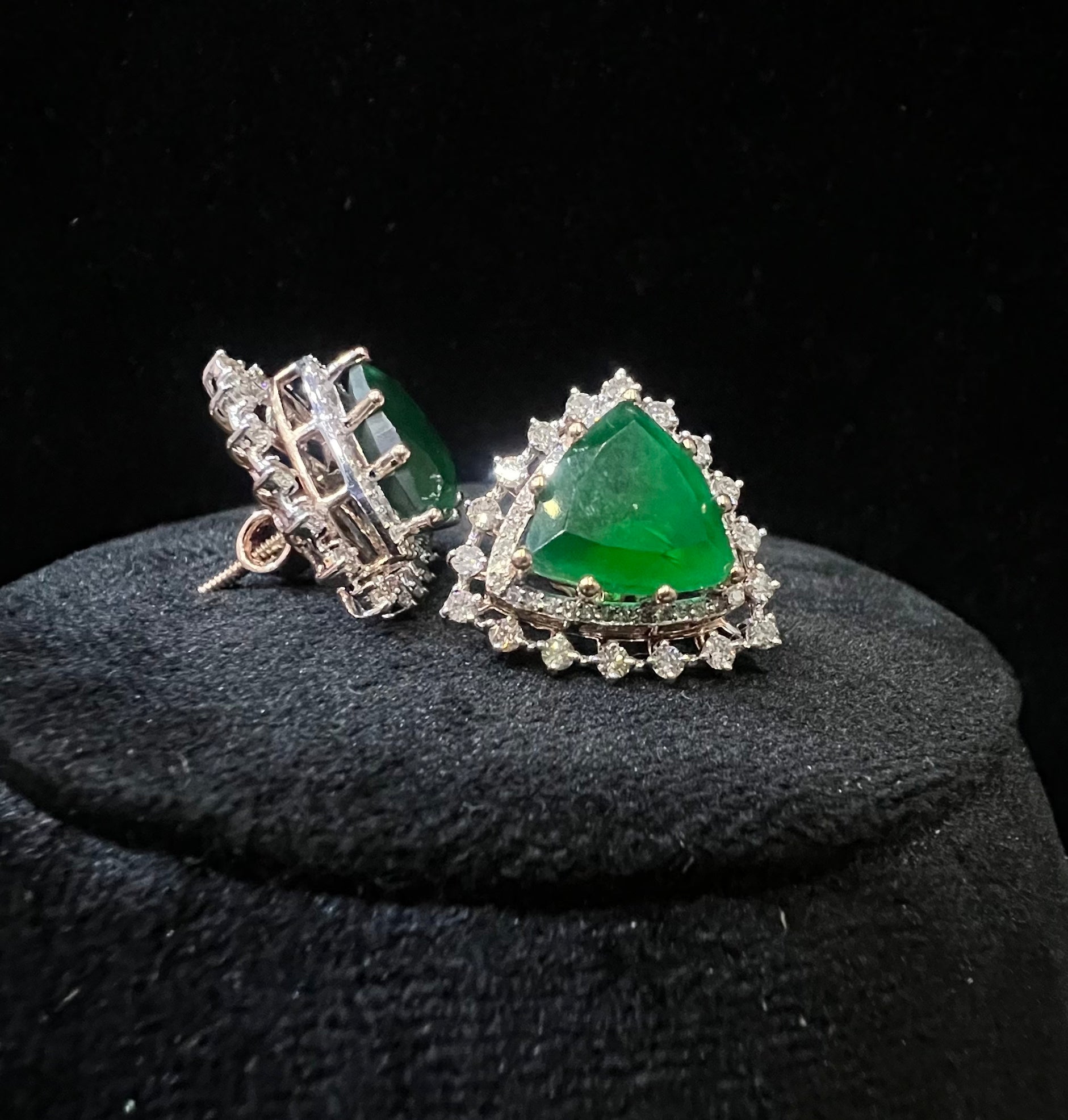 A pair of gold diamond earrings with a green color stone set in a minimalist design on a black background.