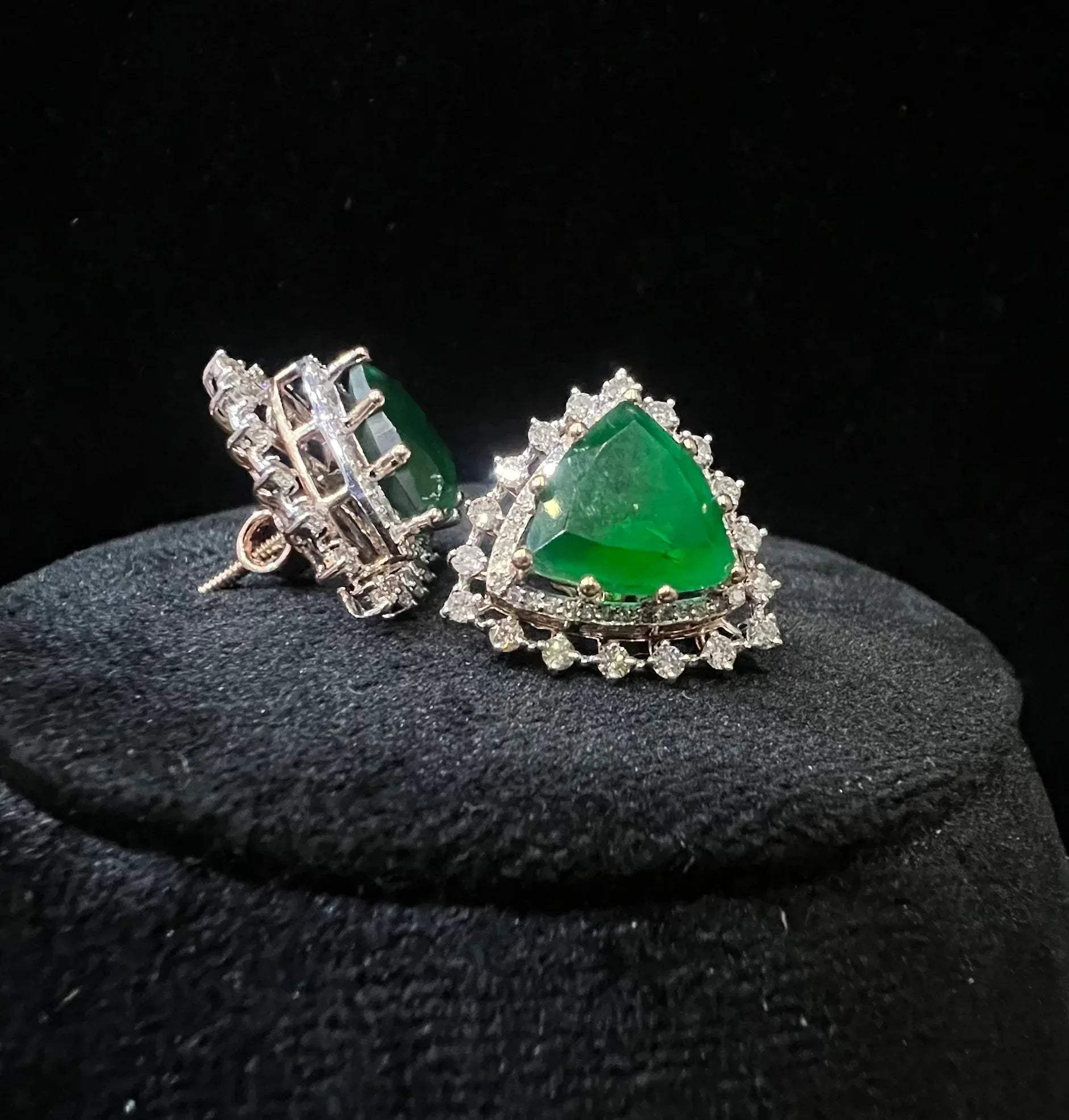 A pair of gold diamond earrings with a green color stone set in a minimalist design on a black background.