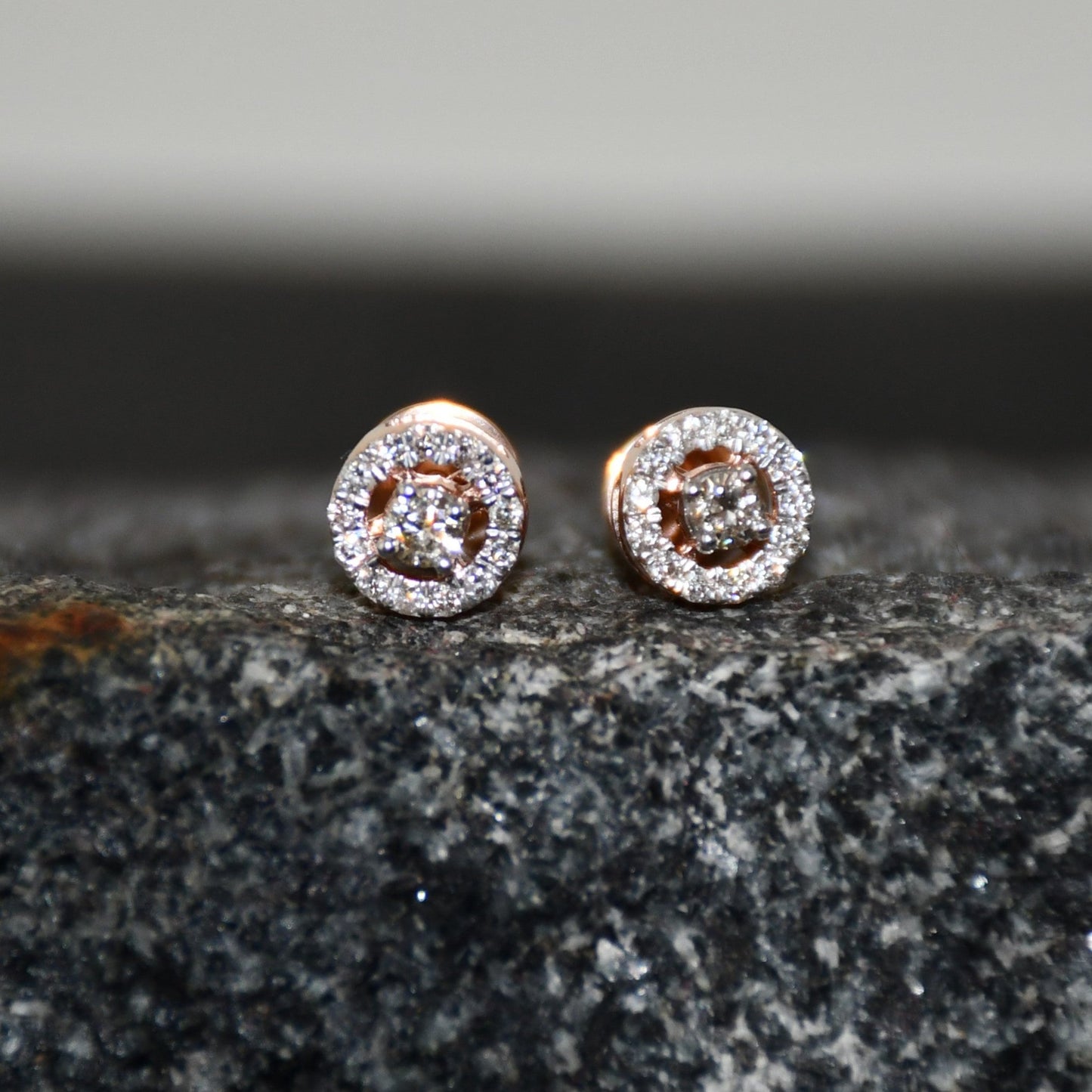 Pair of round halo cluster diamond earrings in gold with center diamonds and surrounding diamond halo, placed on a textured stone surface.