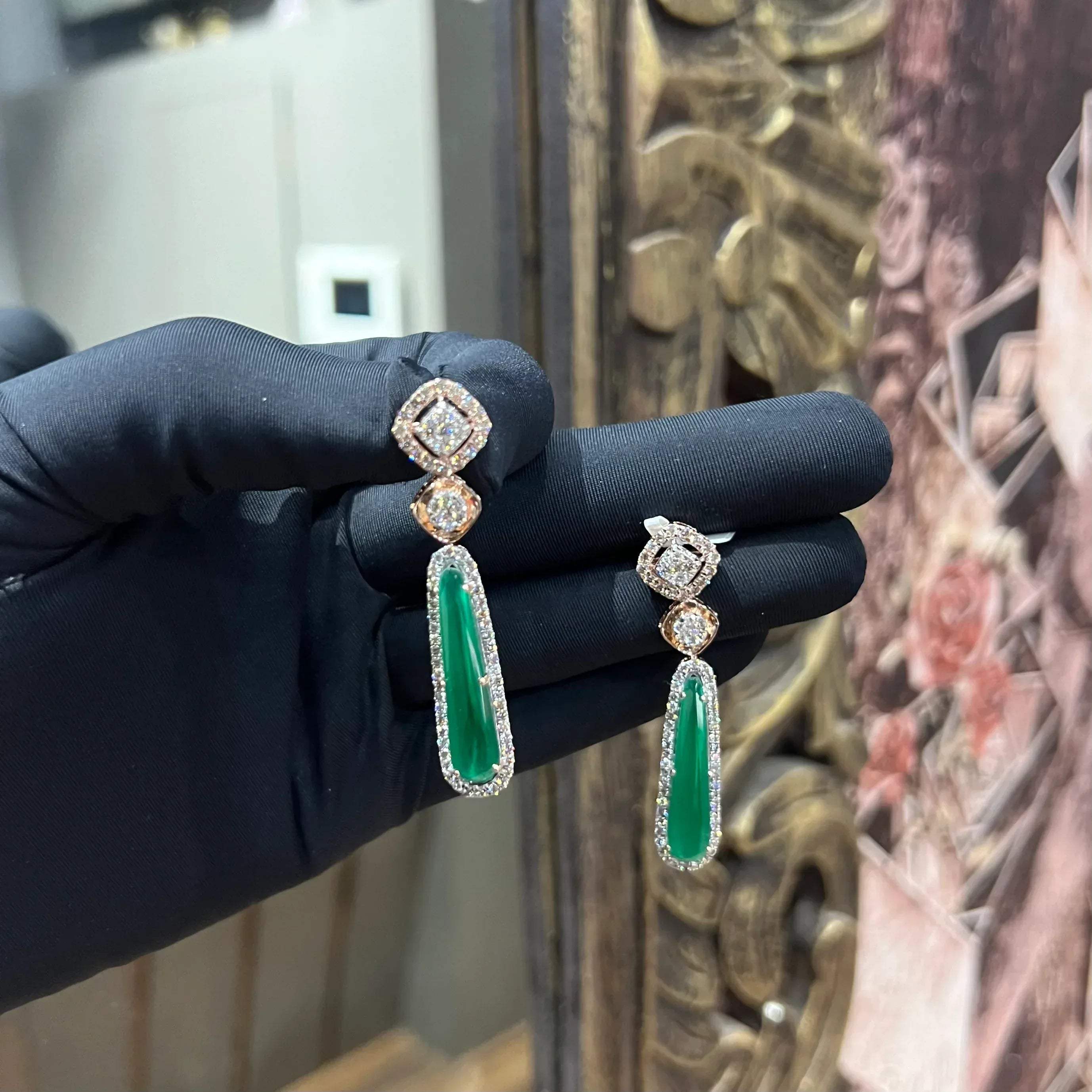 A pair of gold diamond earrings with green color stones set in a minimalist design.