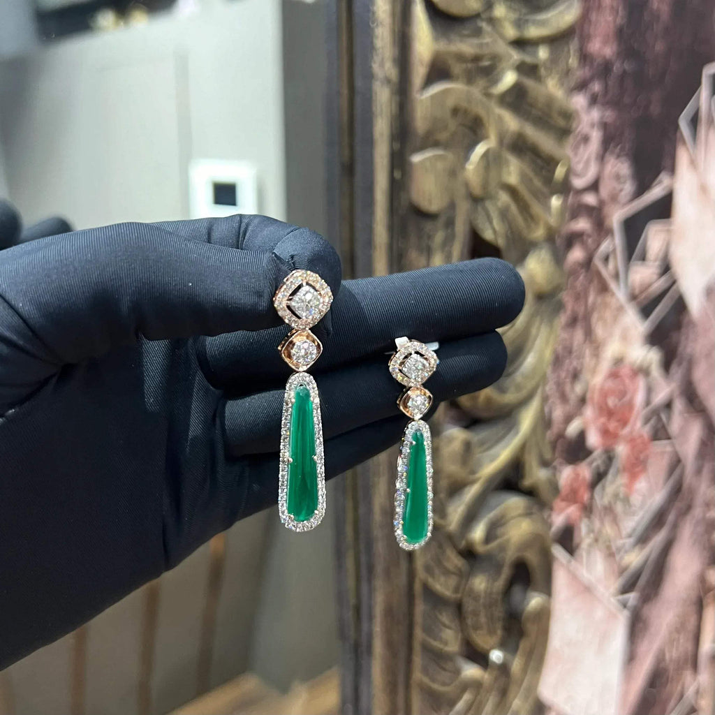 A pair of gold diamond earrings with green color stones set in a minimalist design.