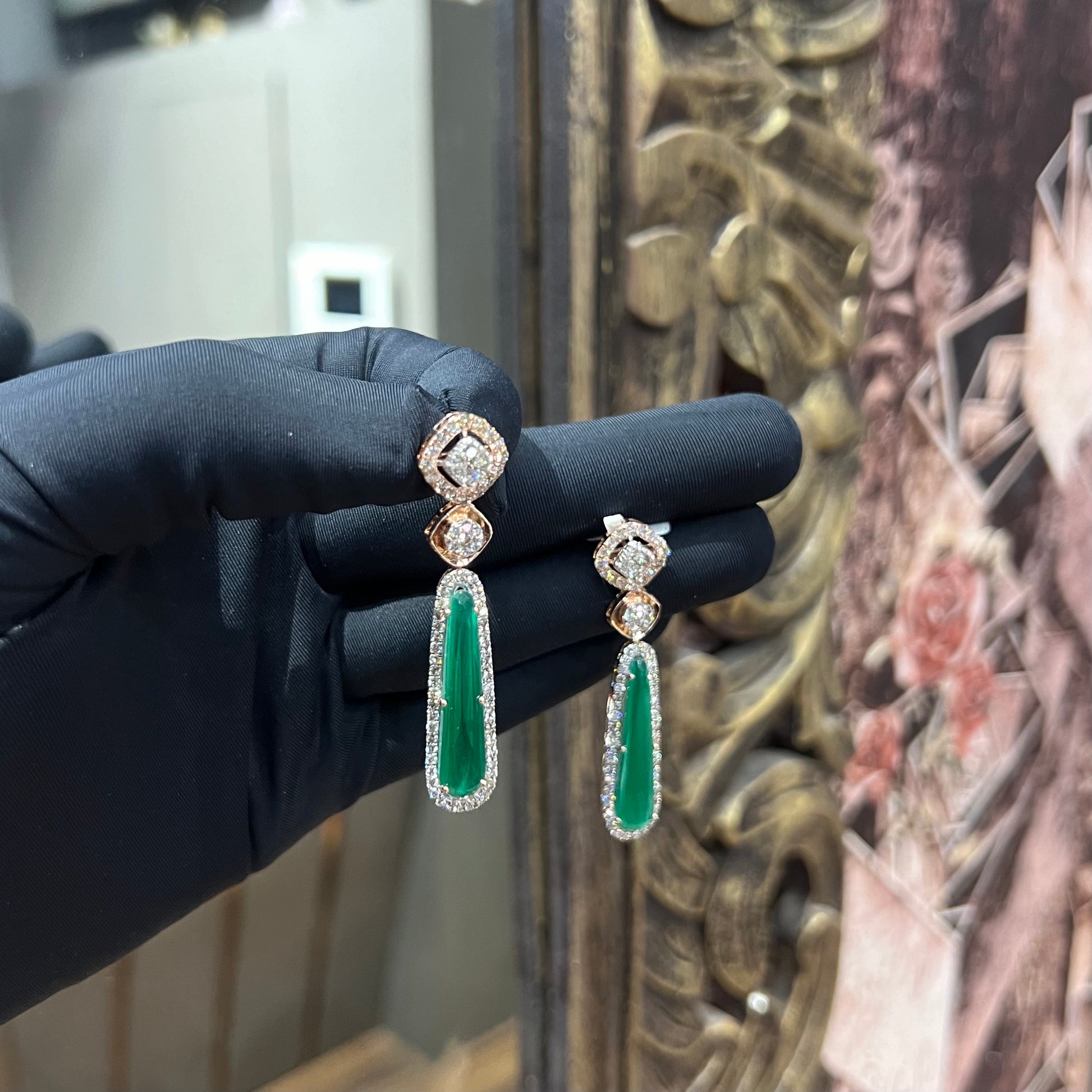 A pair of gold diamond earrings with green color stones set in a minimalist design.