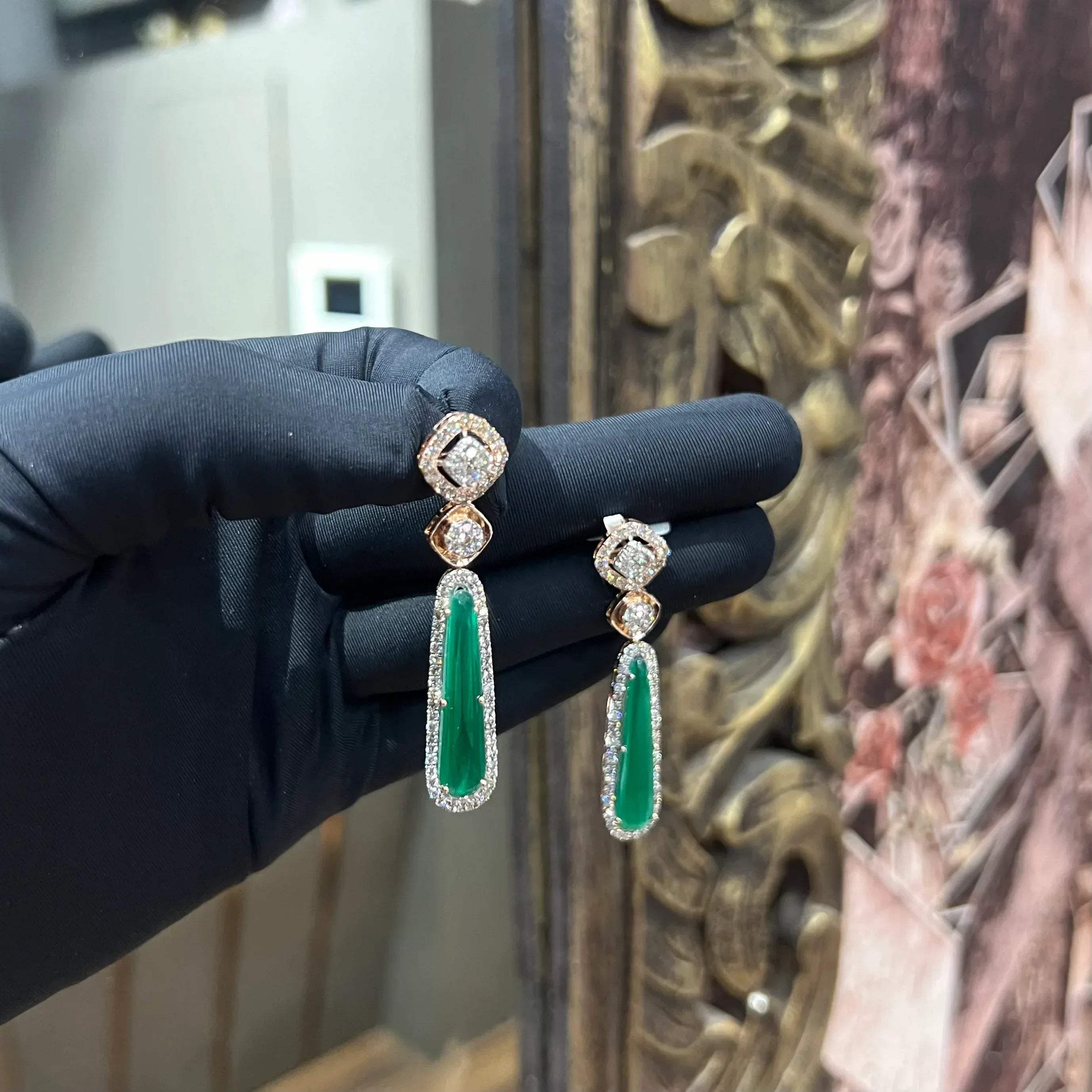 A pair of gold diamond earrings with green color stones set in a minimalist design.