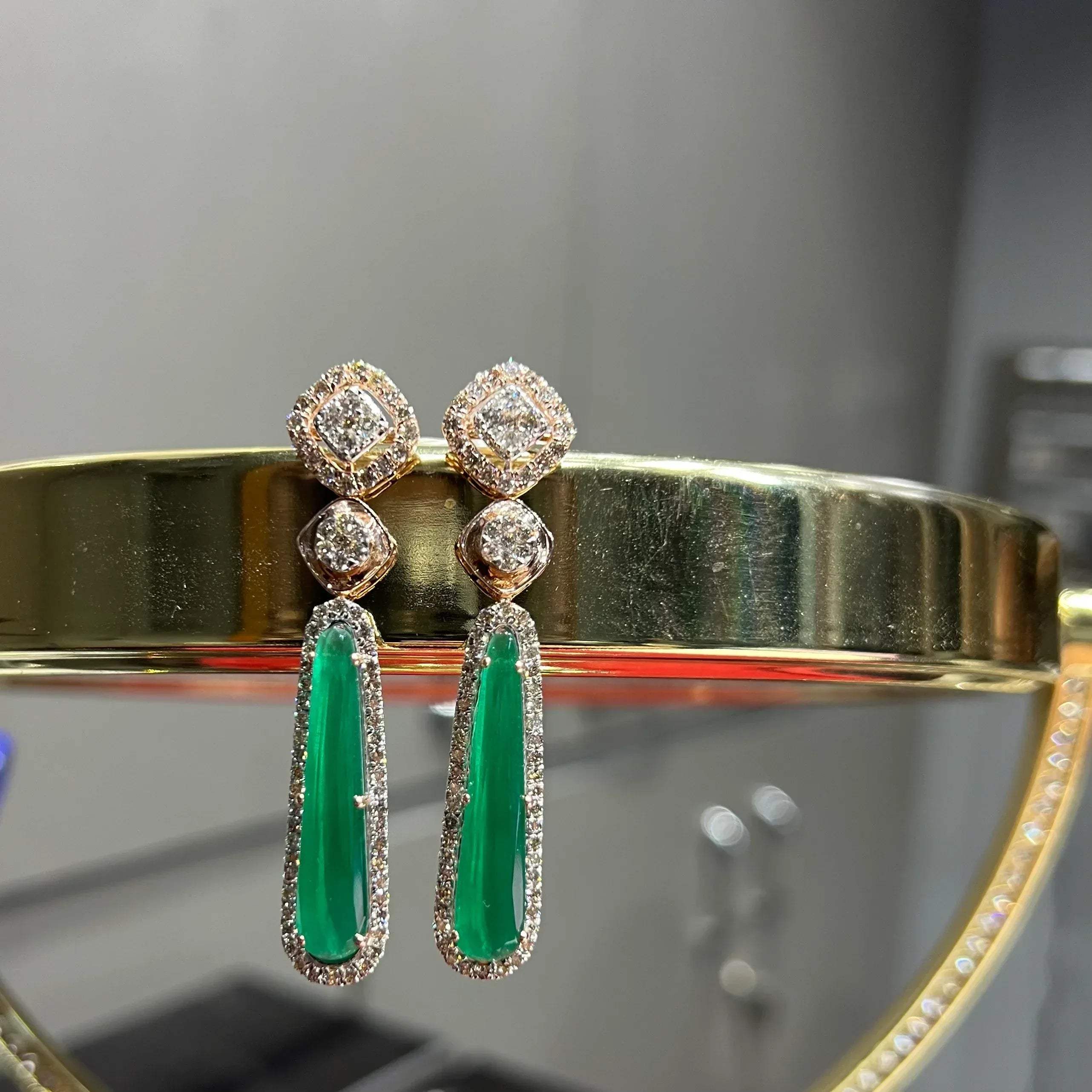 A pair of gold diamond earrings with green color stones set in a minimalist design.