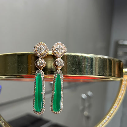 A pair of gold diamond earrings with green color stones set in a minimalist design.