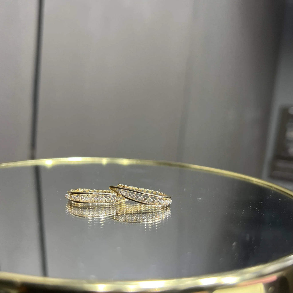 A pair of round-cut diamond hoop earrings held in a person's hand, with a gold band.