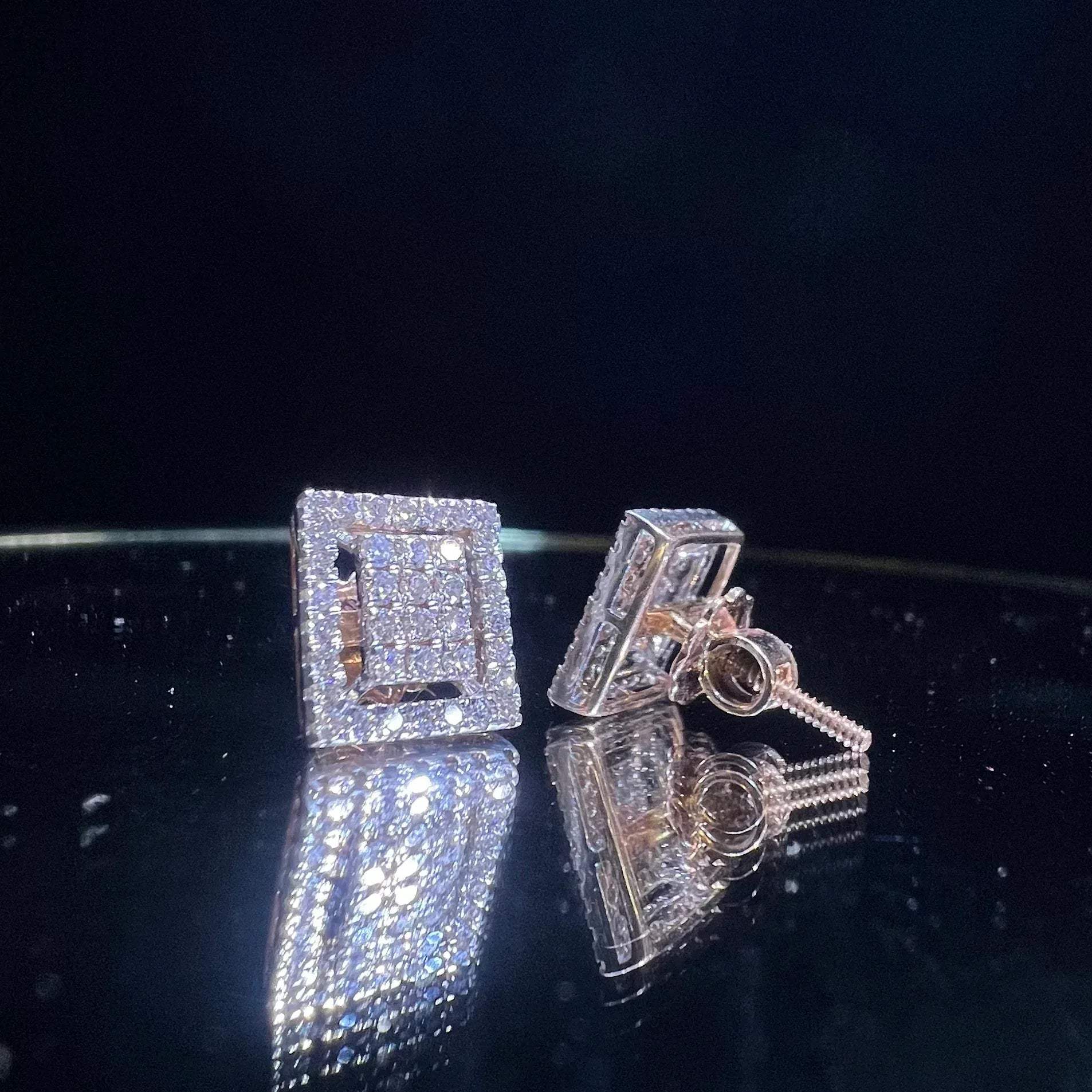 A pair of diamond stud earrings set in a gold casing, displayed against a reflective surface.