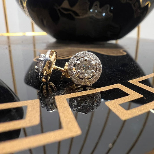 A pair of gold diamond earrings with a round cut design, placed on a reflective surface with a decorative background.