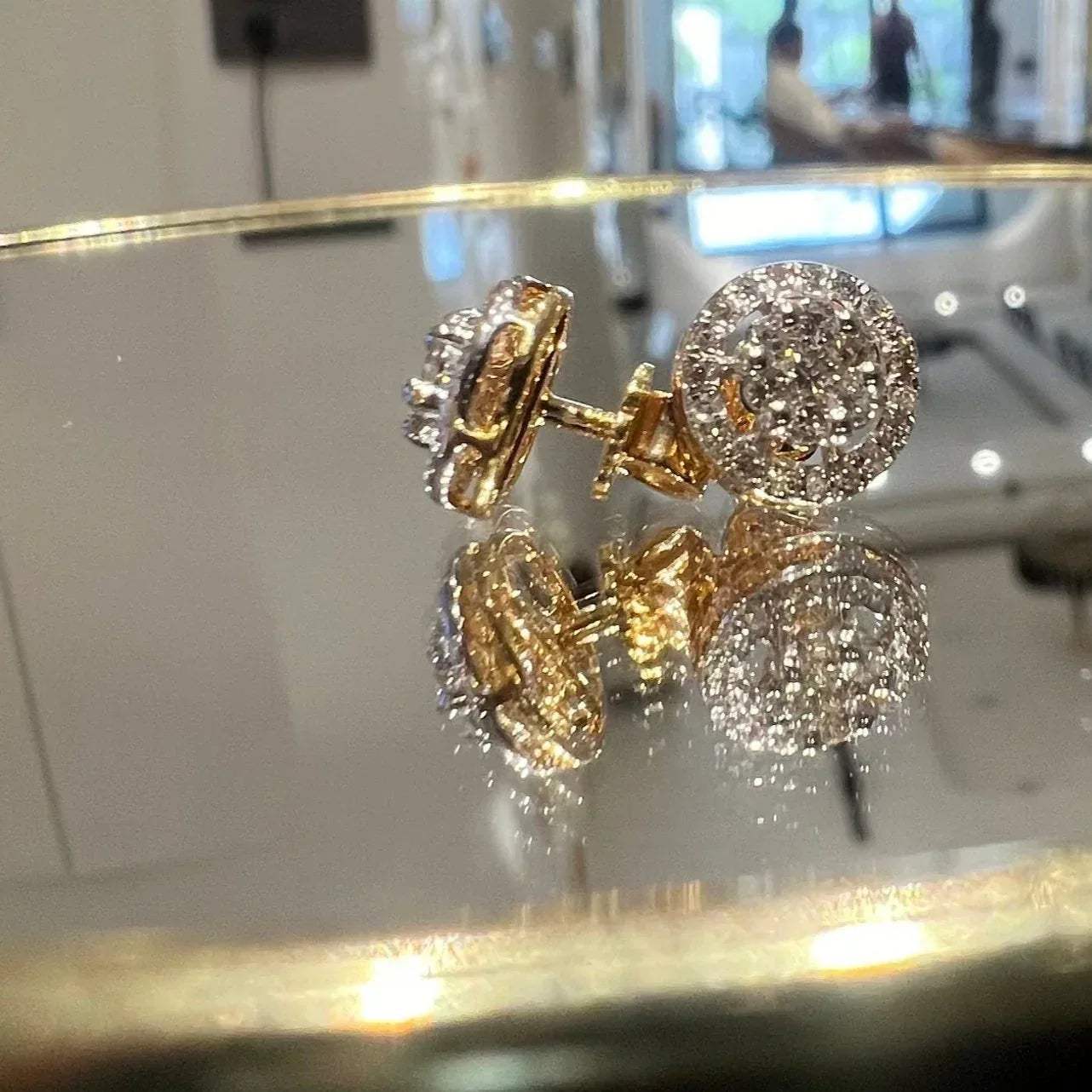 A pair of gold diamond earrings with a round cut design, placed on a reflective surface with a decorative background.