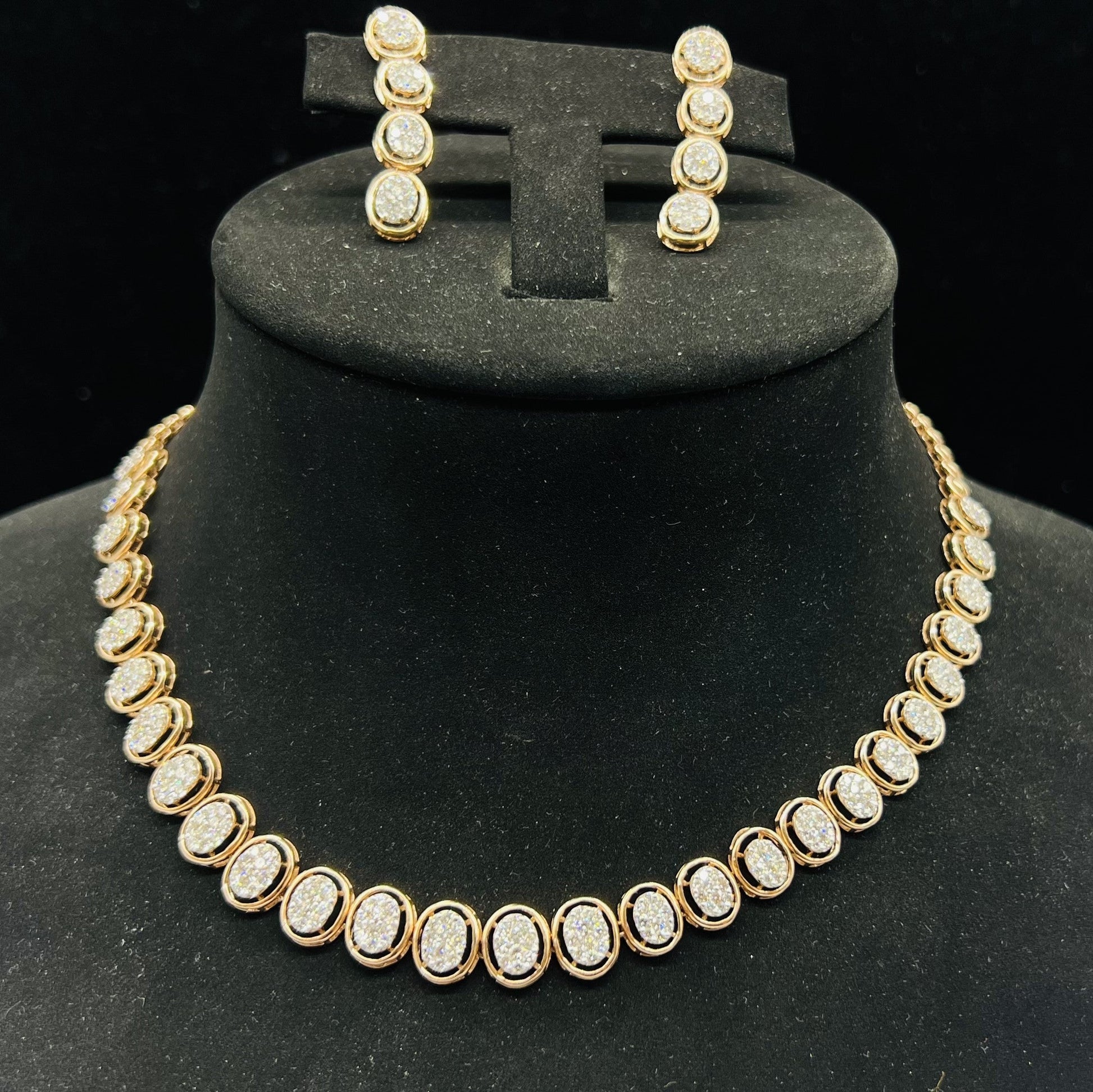 Gold diamond necklace set with matching earrings featuring oval-shaped, pavé-set diamonds in rose gold. The necklace has a linked design showcasing these sparkling stones evenly spaced. Earrings mirror the necklace stones in a vertical stack, creating an elegant accessory set on a black velvet background.