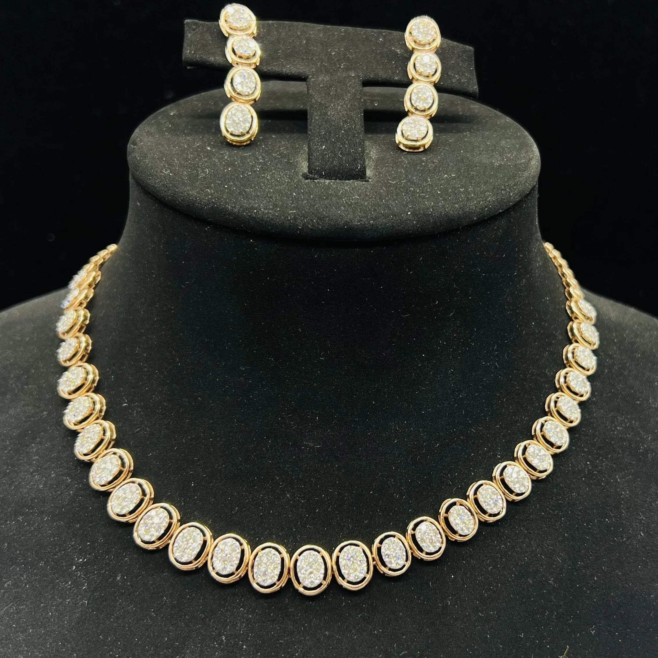 Gold diamond necklace set with matching earrings featuring oval-shaped, pavé-set diamonds in rose gold. The necklace has a linked design showcasing these sparkling stones evenly spaced. Earrings mirror the necklace stones in a vertical stack, creating an elegant accessory set on a black velvet background.