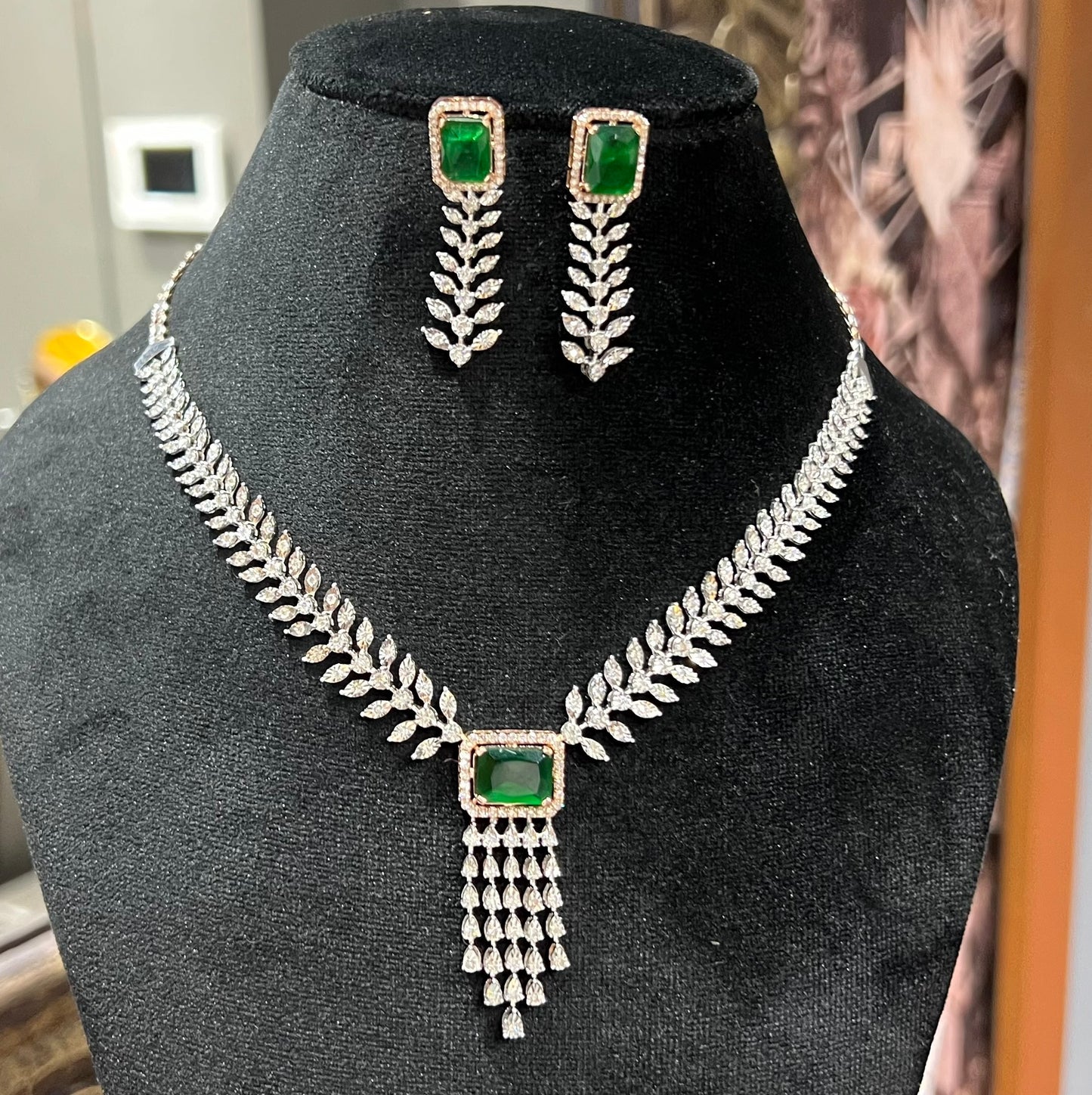 Diamond necklace set featuring a radiant green color stone centerpiece framed by diamond leaves and cascading diamond drops, paired with matching earrings on a mannequin bust.