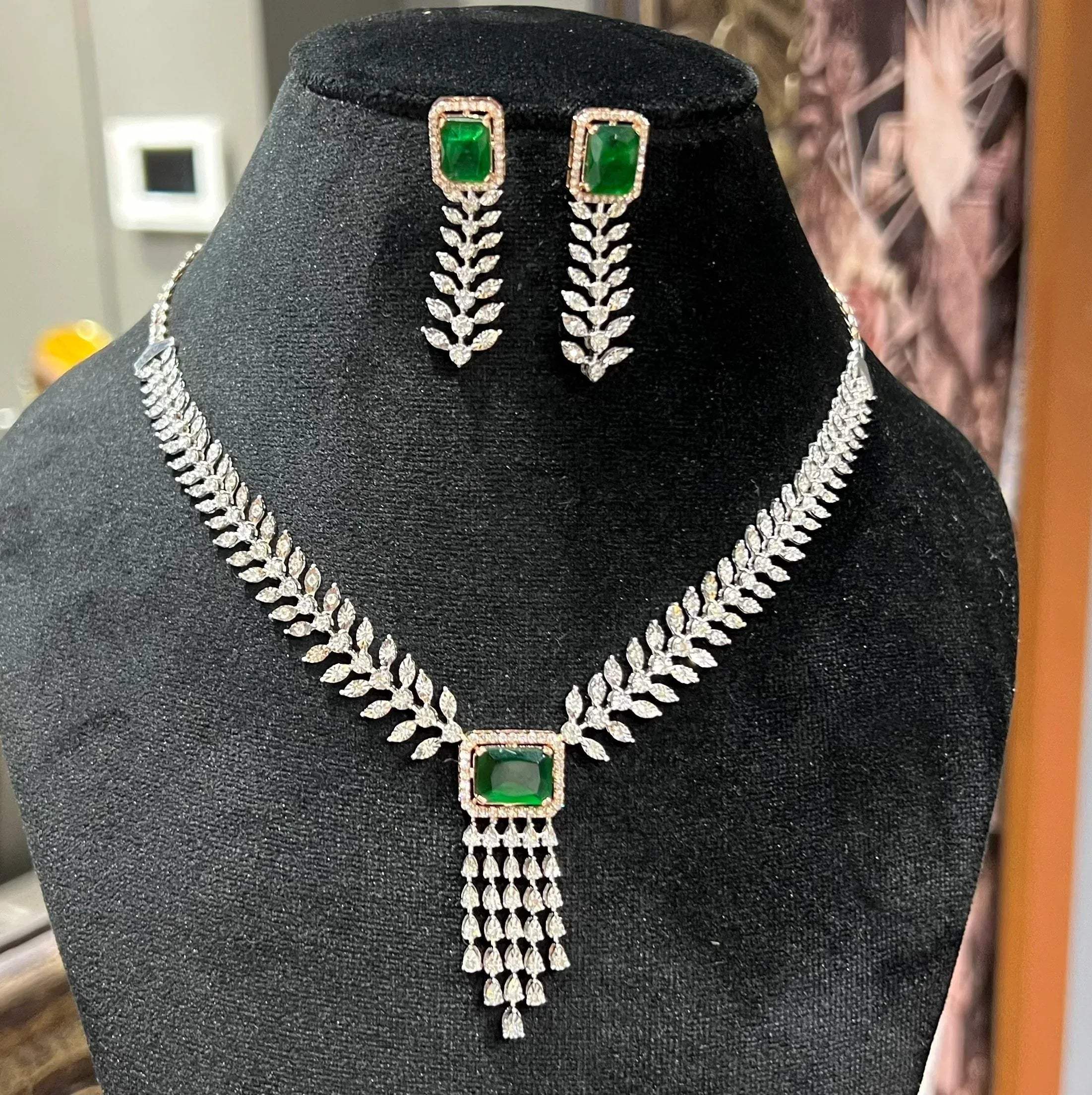 Diamond necklace set featuring a radiant green color stone centerpiece framed by diamond leaves and cascading diamond drops, paired with matching earrings on a mannequin bust.