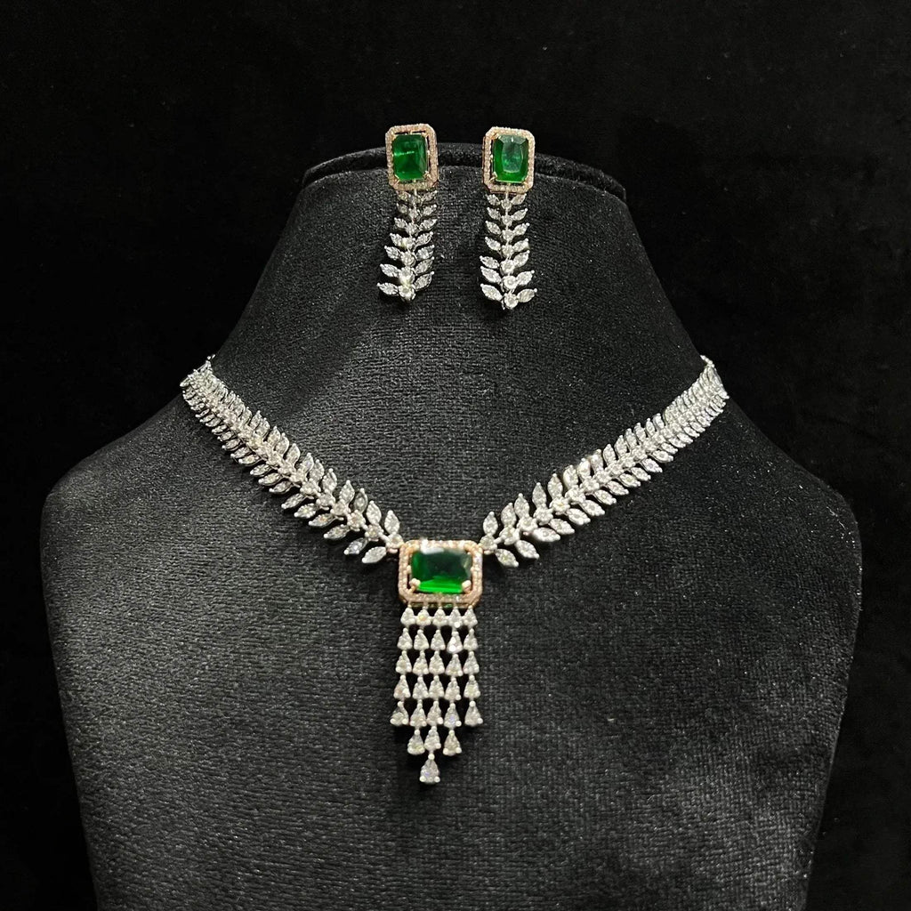 Diamond necklace set featuring a radiant green color stone centerpiece framed by diamond leaves and cascading diamond drops, paired with matching earrings on a mannequin bust.
