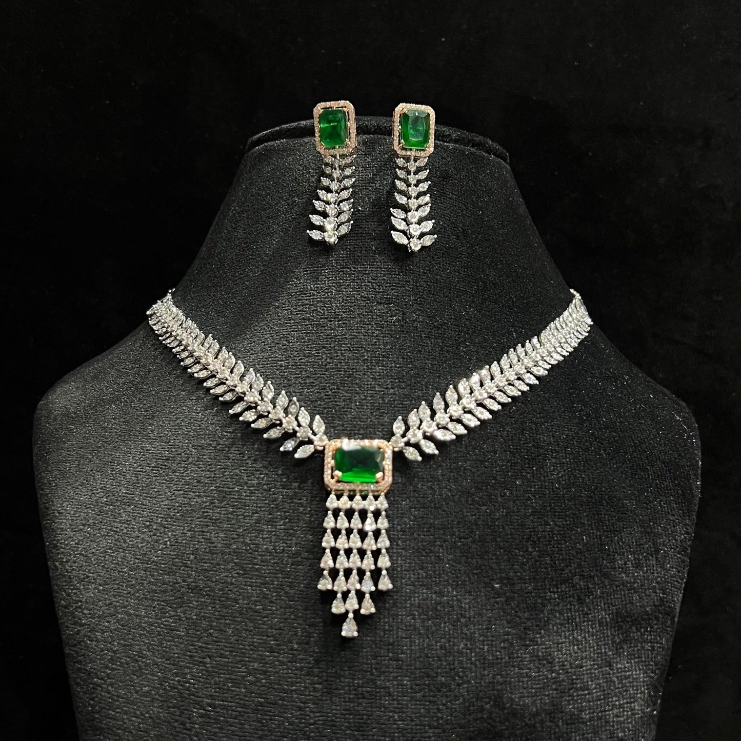 Diamond necklace set featuring a radiant green color stone centerpiece framed by diamond leaves and cascading diamond drops, paired with matching earrings on a mannequin bust.