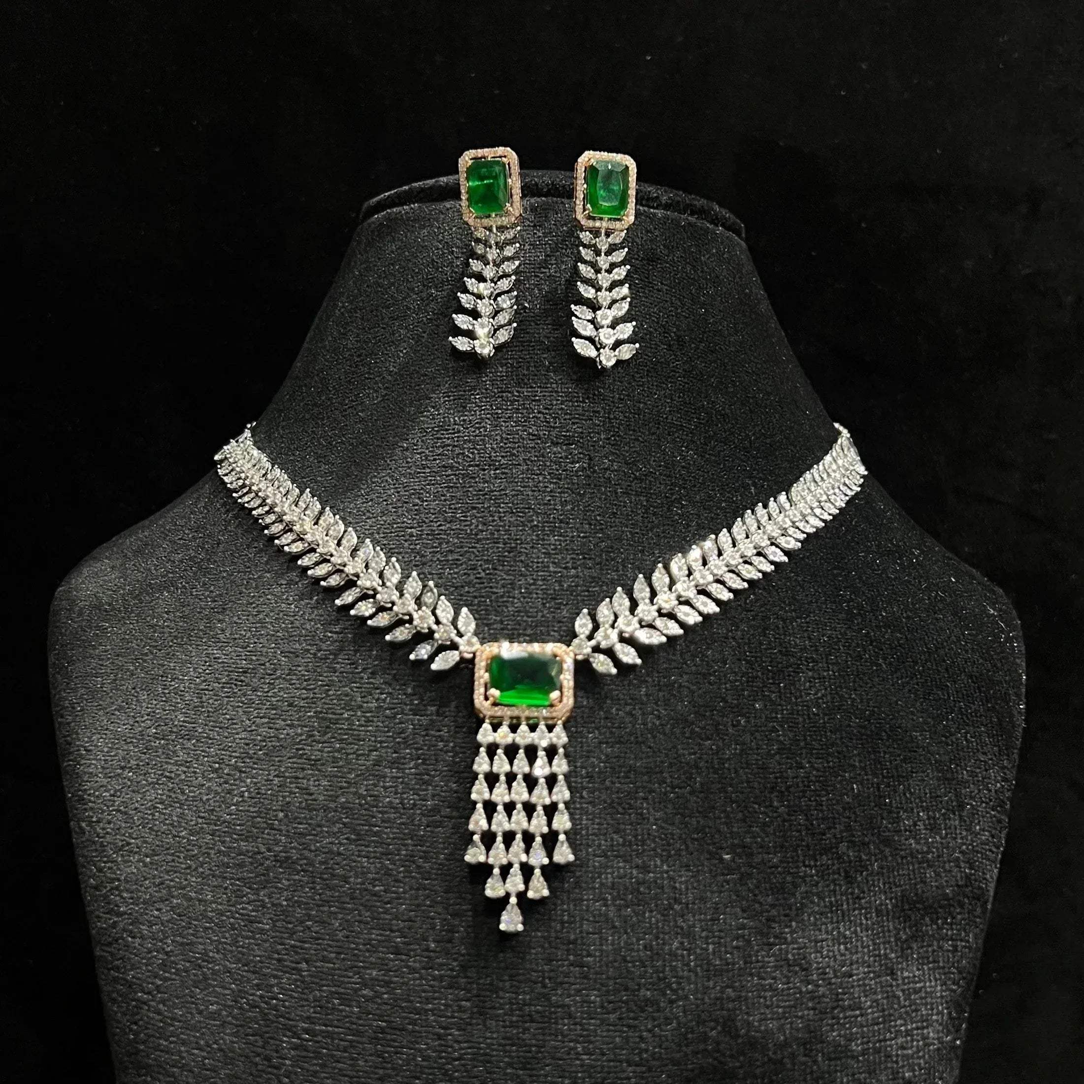 Diamond necklace set featuring a radiant green color stone centerpiece framed by diamond leaves and cascading diamond drops, paired with matching earrings on a mannequin bust.