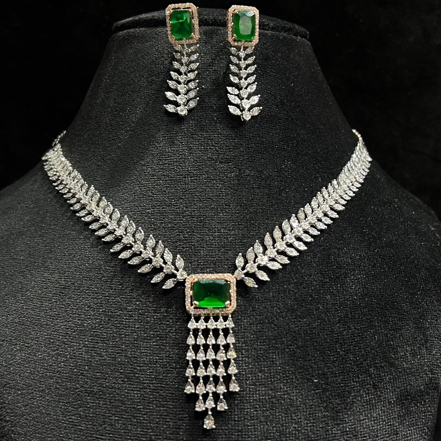 Diamond necklace set featuring a radiant green color stone centerpiece framed by diamond leaves and cascading diamond drops, paired with matching earrings on a mannequin bust.