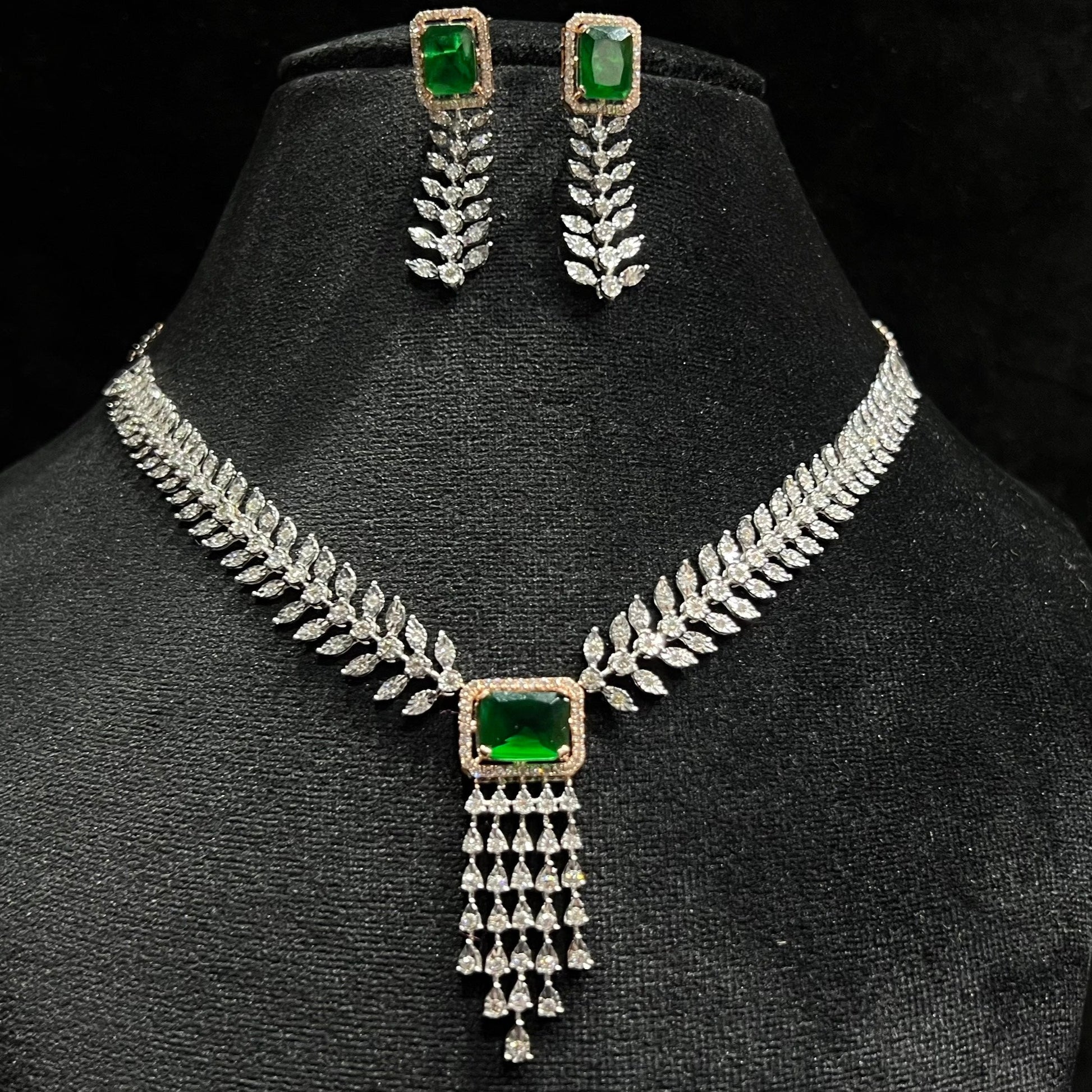 Diamond necklace set featuring a radiant green color stone centerpiece framed by diamond leaves and cascading diamond drops, paired with matching earrings on a mannequin bust.