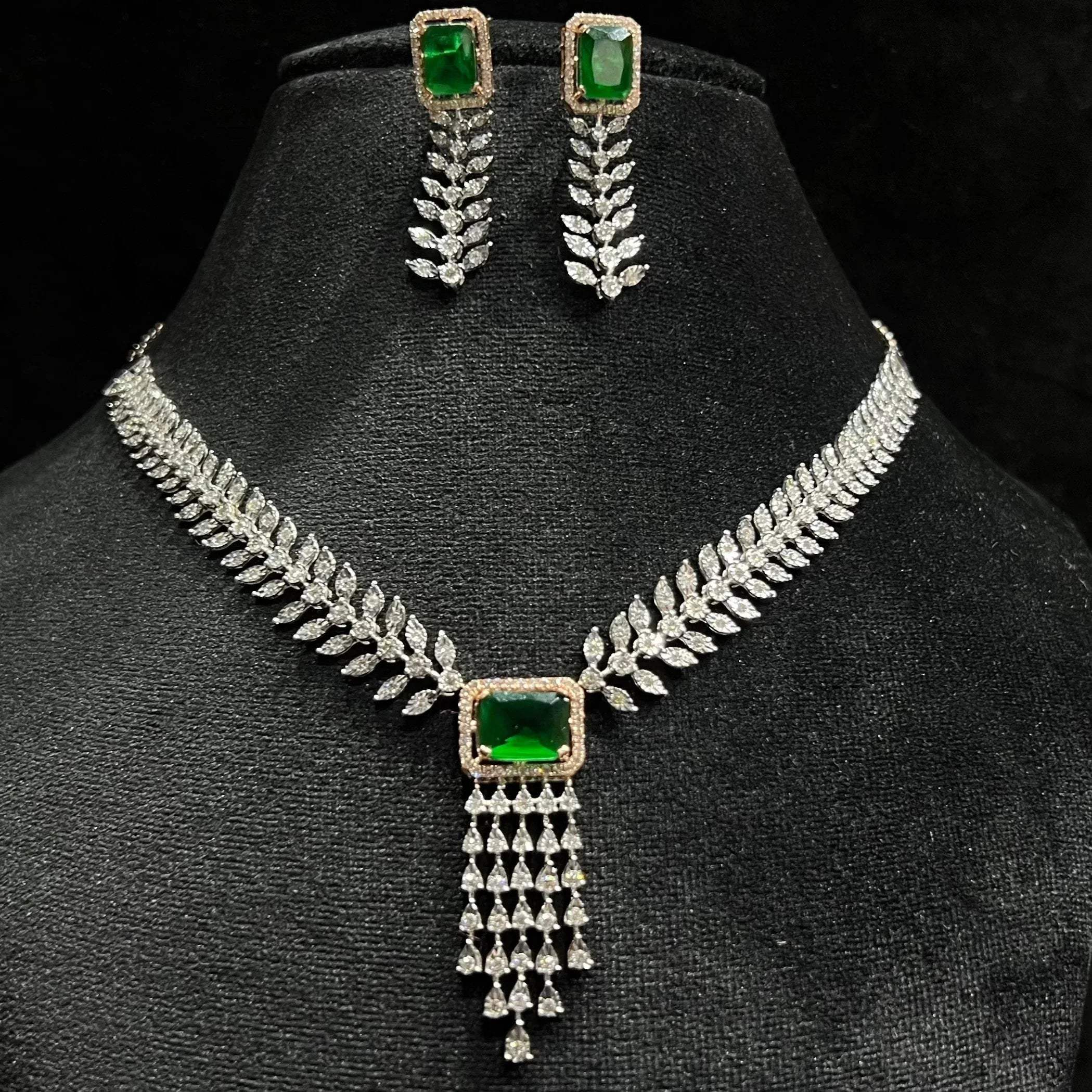 Diamond necklace set featuring a radiant green color stone centerpiece framed by diamond leaves and cascading diamond drops, paired with matching earrings on a mannequin bust.